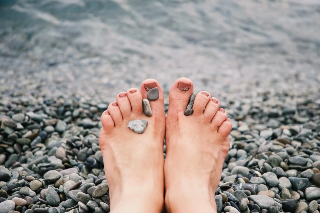 Eine Junge Frau die gerne Barfußschuhe trägt, streckt ihre beiden Füße an einem Steinstrand aus.